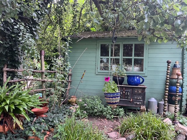 Eine grüne Hütte umgeben von Pflanzen, Töpfen und Dekorativem im Garten.