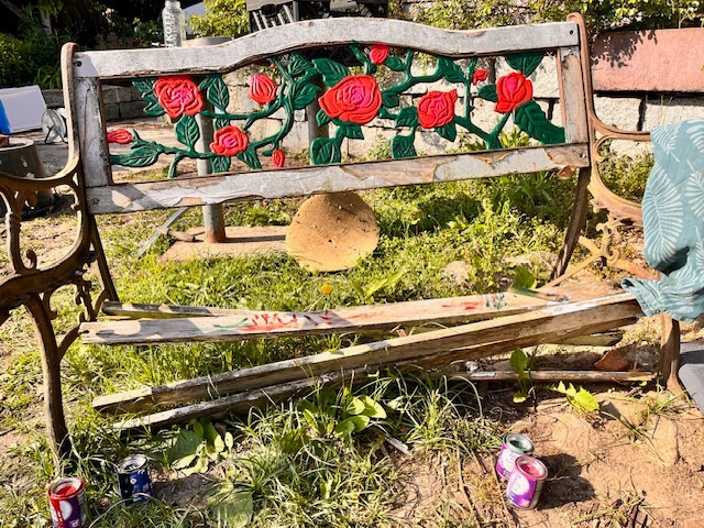 Eine beschädigte Gartenbank mit roten Rosenmotiven und rostigem Metallrahmen, umgeben von Gras und leeren Dosen.