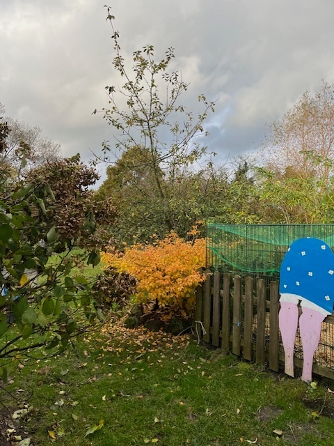 Ein Garten mit bunten Pflanzen und einem hölzernen Zaun, im Hintergrund ein strahlender Baum und ein großer, blau bemalter Mosaik mit einem orangenen Unterteil und rosa Beinen.