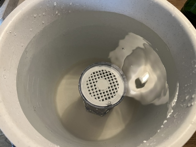Ein weißer Eimer mit Wasser, in dessen Mitte ein durchsichtiger Filterbehälter steht.