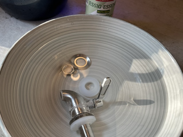 Eine metallene Armatur und verschiedene Dichtungen befinden sich in einer weißen Schüssel mit Wasser, im Hintergrund ein Flasche Essig.