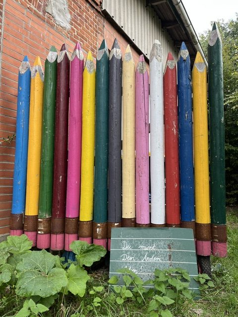 Ein bunter Stift-Ständer aus Holz mit großen, dekorativen Buntstiften in verschiedenen Farben, platziert vor einer Backsteinmauer.
