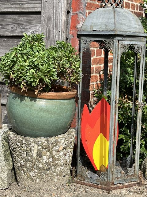 Blumentopf mit grünen Pflanzen aus Keramik neben einer antiken Laterne mit buntem Holzschild in Form einer Blume.