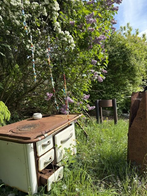 Ein verrosteter alter Ofen steht in einem üppigen Garten, umgeben von blühenden Sträuchern und grünem Gras. Im Hintergrund sind ein Holzstuhl und weitere Pflanzen zu sehen.