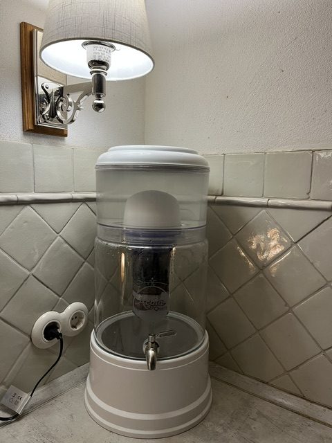 Ein Wasserspender mit transparentem Behälter und Hahn, neben einer Wandlampe in einem Raum mit weißen Fliesen.