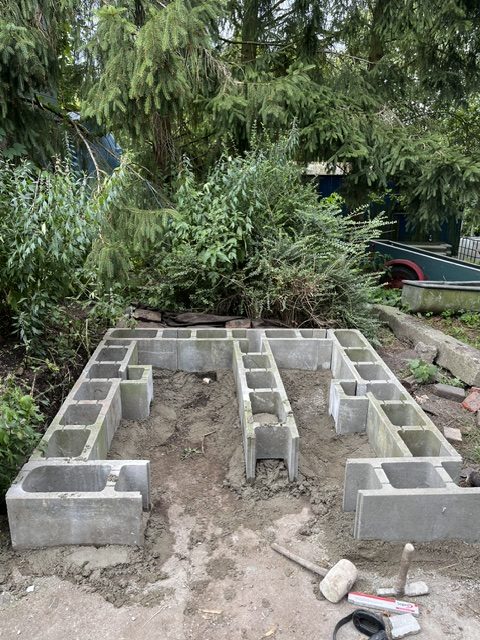 Ein unvollendeter Mauerwerksblockrahmen im Garten, umgeben von Büschen und Bäumen.