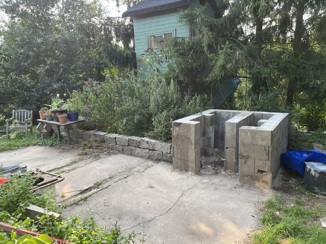 Blick auf einen Garten mit einer unvollendeten Mauer aus Betonsteinen, einer kleinen Gartenhütte und einem bewachsenen Bereich mit Blumentöpfen.
