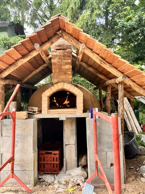 Ein traditioneller Holzofen mit einem steinernen Kamin, umgeben von einem Holzdach und Baugerüsten, in einer grünen Umgebung.