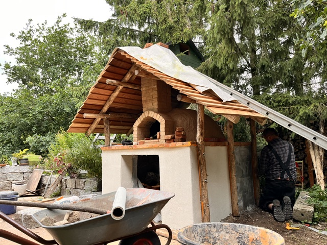 Ein Gartenofen mit einem Holzdach, umgeben von Bäumen und Pflanzen. Im Vordergrund ein Schubkarren und eine Person, die am Ofen arbeitet.