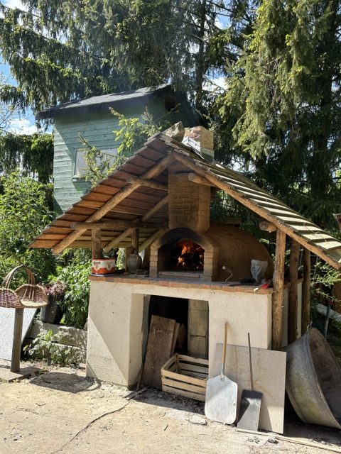 Ein Backofen im Freien mit einem Holzdach und einer Feuerstelle, umgeben von Bäumen und Pflanzen.