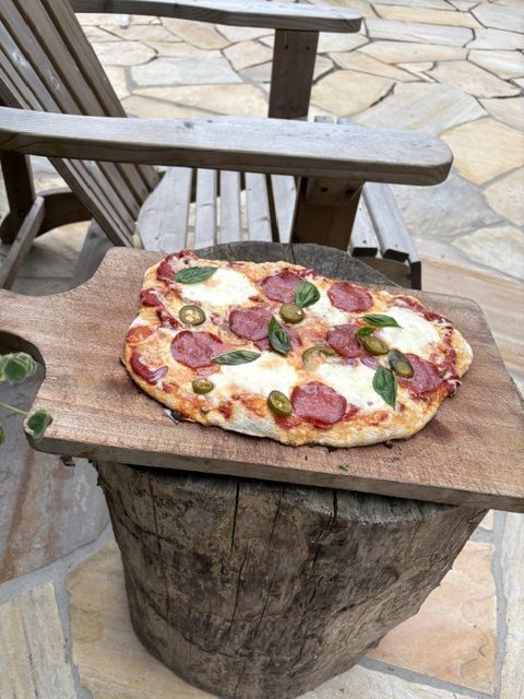 Ein Holzbrett mit einer Pizza belegt mit Salami, Käse, Jalapeños und frischem Basilikum, platziert auf einem Baumscheit im Freien.