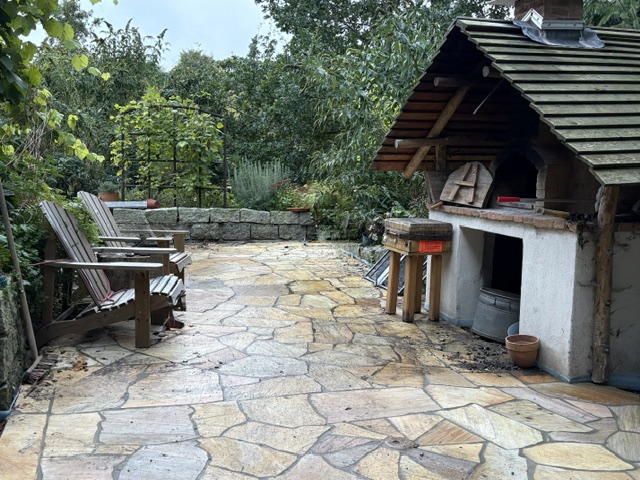 Eine gemütliche Terrasse mit einem gemütlichen Sitzbereich aus Holzstühlen und einem Grillplatz, umgeben von üppigem Grün und Bäumen.